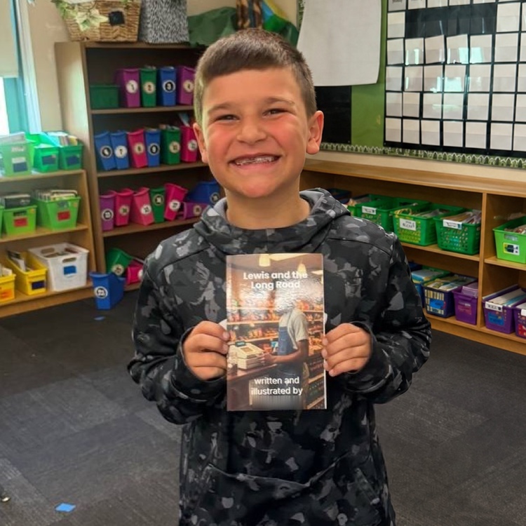 A smiling young author holding a printed copy of his book in a classroom